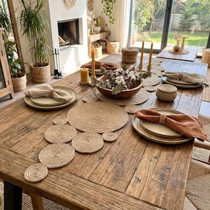 Wide farmhouse table with a jute coil runner, a wooden bowl of cotton stems and eucalyptus, rust linen napkins and beeswax candles by an open fireplace