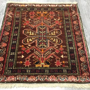 May include: A traditional rug with a dark red background and intricate geometric patterns in shades of red, orange, yellow, green, and black. The rug has a fringe border.