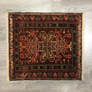 May include: A traditional Persian rug with a dark red background and intricate geometric patterns in shades of red, orange, black, and white. The rug has a fringe border.