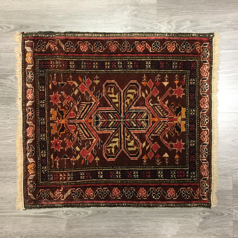 May include: A traditional Persian rug with a dark red background and intricate geometric patterns in shades of red, orange, black, and white. The rug has a fringe border.