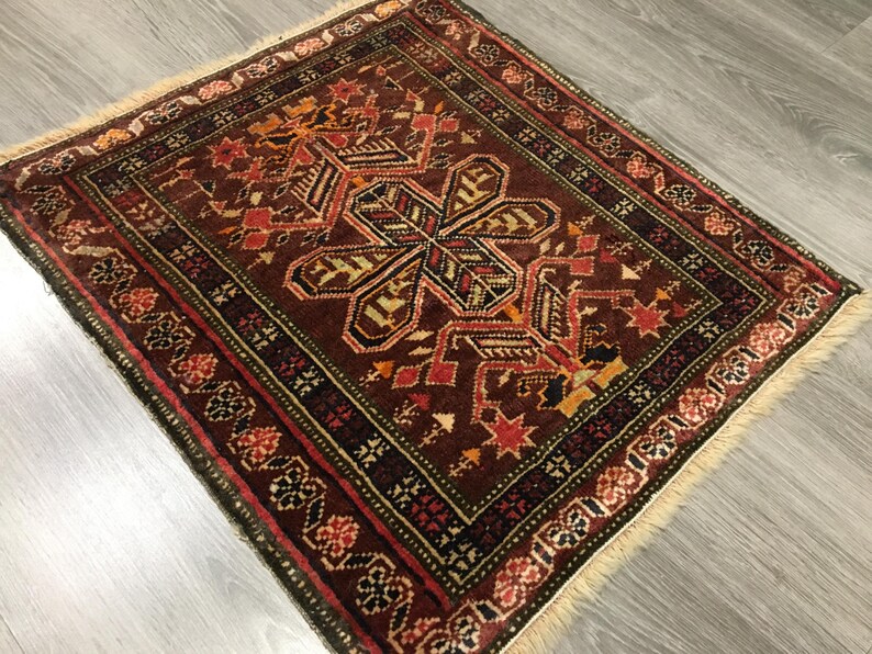 May include: A traditional rug with a geometric pattern in shades of red, brown, green, and cream. The rug has a fringe border and is made of wool.