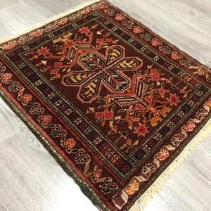 May include: A small, square rug with a traditional Persian design. The rug has a dark brown background with intricate patterns in shades of red, orange, green, and black. The rug has a fringe border.