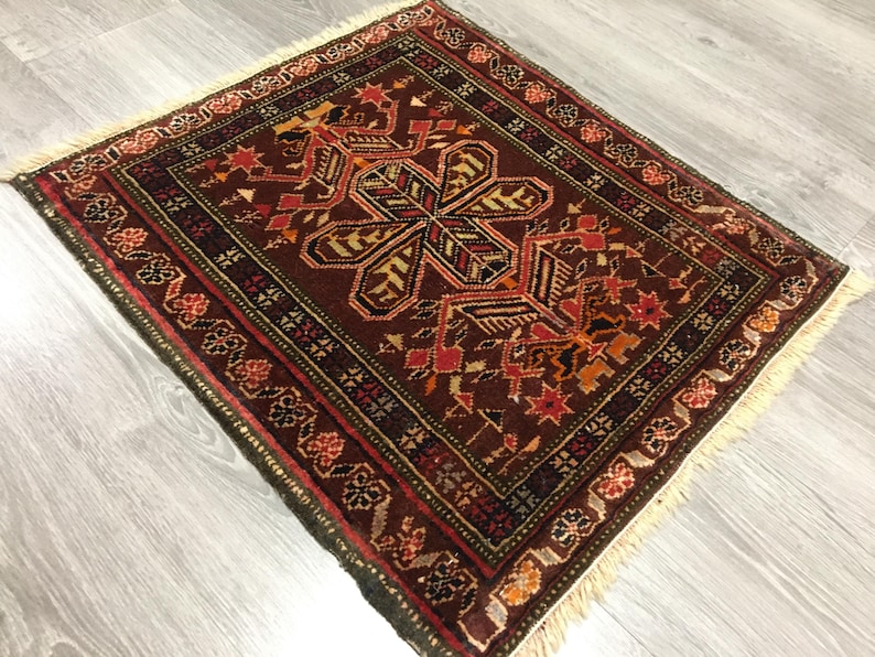 May include: A small, square rug with a traditional Persian design. The rug has a dark brown background with intricate patterns in shades of red, orange, green, and black. The rug has a fringe border.