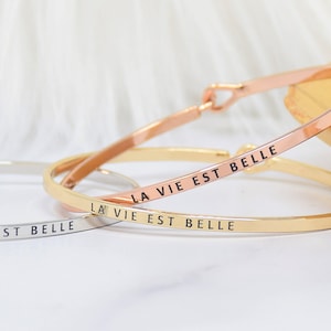 May include: Three silver bangle bracelets with the inscription "La Vie Est Belle" on each. The bracelets are stacked on top of each other.