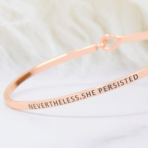 May include: A rose gold bangle bracelet with the inscription "NEVERTHELESS, SHE PERSISTED".