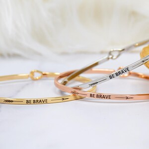 May include: Three thin gold, silver, and rose gold bracelets with the words "Be Brave" and an arrow engraved on each.