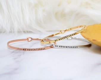 May include: Three gold, silver, and rose gold bangle bracelets with the inscription "BE ADVENTUROUS" on each.