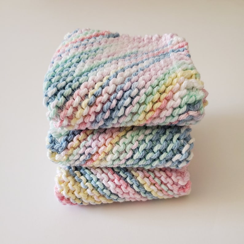 May include: Three knitted dishcloths in a stack. The dishcloths are made with a multi-colored yarn in shades of pink, blue, yellow, and white.