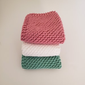 Puede incluir: Tres paños de cocina de punto en rosa, blanco y verde. Los paños están apilados uno encima del otro.
