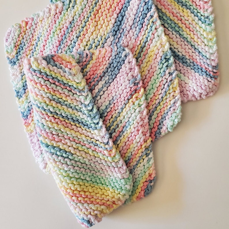 May include: Three colorful striped knitted dishcloths in shades of pink, blue, yellow, and green. The dishcloths are folded on top of each other.