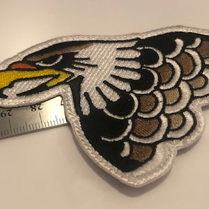 Può includere: Toppa ricamata bianca con la testa di un'aquila marrone e nera con un becco giallo. La testa dell'aquila è rivolta a sinistra e le penne dell'ala sono stilizzate.