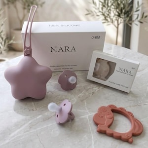 Op de afbeelding: Een verzameling babyproducten in mauve en perziktinten, waaronder een ster-vormige fopspeenhouder, fopspenen en een lam-vormige bijtring. De verpakking toont de tekst "100% SILICONE", "NARA" en "0-6M". De bijtring is gelabeld met "SILICONE LAMB TEETHER".