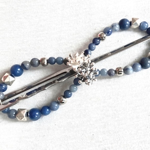 Puede incluir: Una horquilla de metal plateado con un marco de alambre en forma de infinito adornado con una variedad de cuentas azules, esferas plateadas lisas y pequeños tachones piramidales plateados, centrada con un dije plateado en forma de flor o estrella.