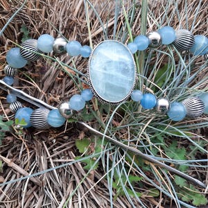 Puede incluir: Un accesorio para el cabello con una gran piedra cabujón ovalada de color azul claro en un marco plateado. La pieza está adornada con cuentas azul claro alternadas con cuentas metálicas plateadas, incluyendo diseños esféricos lisos y estriados.