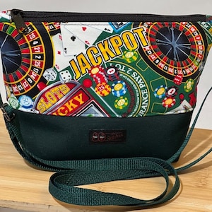 May include: A green fabric purse with a black zipper and a shoulder strap. The purse has a casino themed print with a roulette wheel, playing cards, dice, and the word "JACKPOT".
