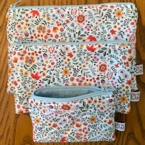 May include: A set of three zippered pouches in varying sizes, featuring a white floral pattern with red, orange, yellow, and blue flowers. Each pouch has a light blue zipper and trim. The pouches are made of fabric.