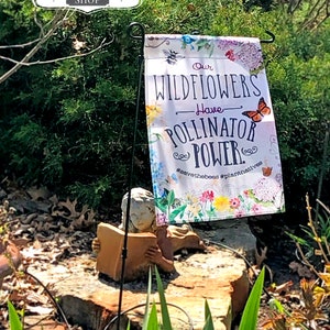 May include: A white garden flag with a floral design and the text "Our Wildflowers Have Pollinator Power. #savethebees #plantnatives".