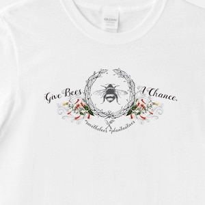 May include: White t-shirt with a black and white bee illustration surrounded by a wreath of flowers and leaves. The text "Give Bees A Chance." is written above the bee and "savethebees plantualives" is written below the bee.