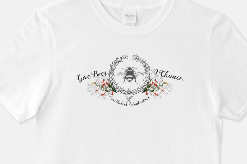 May include: White t-shirt with a black and white bee illustration surrounded by a wreath of flowers and leaves. The text "Give Bees A Chance." is written above the bee and "savethebees plantualives" is written below the bee.
