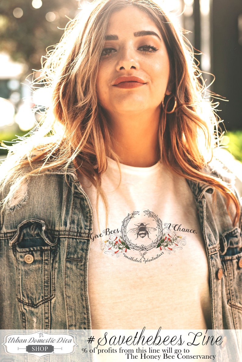 May include: A white t-shirt with a graphic of a bee in a wreath with the text "Give Bees A Chance." and "savethebees" plantualnes. The shirt also has the text "#Savethebees Line" and "% of profits from this line will go to The Honey Bee Conservancy."  The shirt is being worn by a person with long blonde hair and a denim jacket.