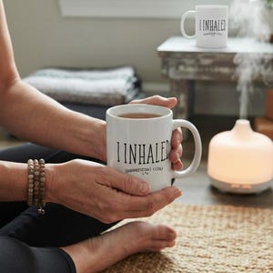 May include: A white ceramic mug with a handle, featuring the black text "I INHALED" and "(essential oils)" printed on the front. A second identical mug is visible in the background, along with a glowing white essential oil diffuser emitting steam.