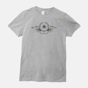 May include: A heather grey t-shirt with a bee and floral wreath design. The text "Give Bees A Chance" is written above the wreath and "pollinators plant lovers" is written below the wreath.