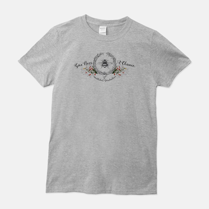 May include: A heather grey t-shirt with a bee and floral wreath design. The text "Give Bees A Chance" is written above the wreath and "pollinators plant lovers" is written below the wreath.