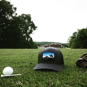May include: A black and blue baseball cap with a skyline and a person riding a horse on the front. The cap is on a green grassy field with a golf ball and tee in the foreground.