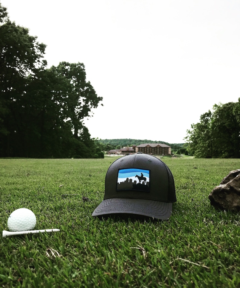 May include: A black and blue baseball cap with a skyline and a person riding a horse on the front. The cap is on a green grassy field with a golf ball and tee in the foreground.