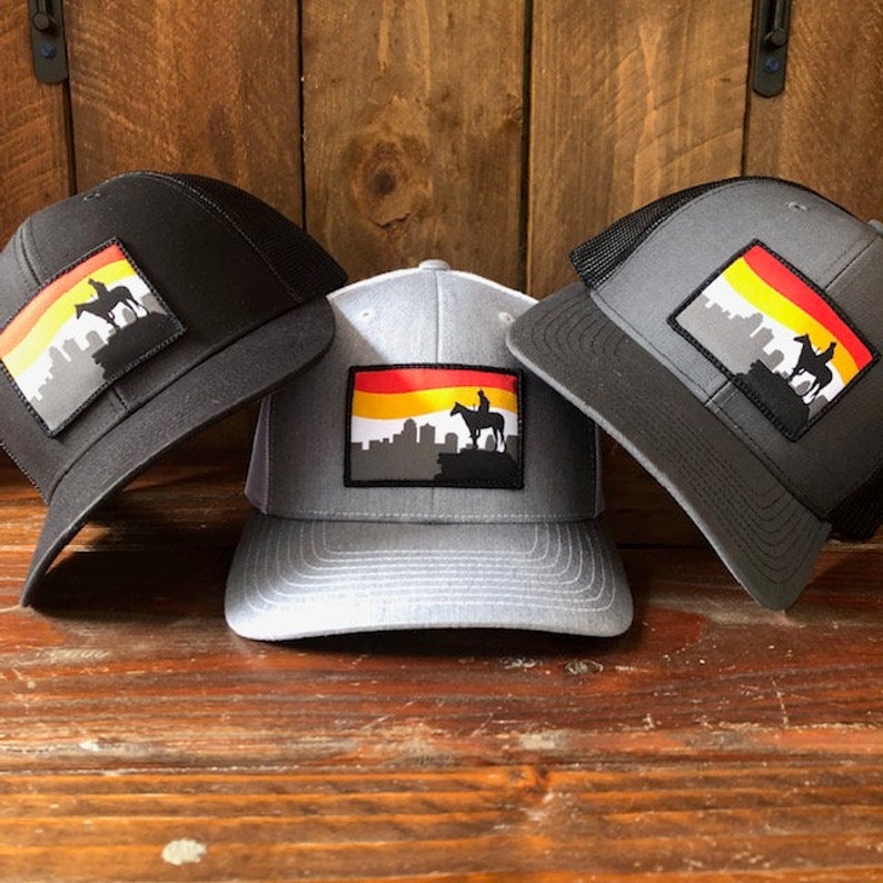 May include: Three trucker hats with a black, gray, and white mesh back. Each hat features a black and white patch with a silhouette of a city skyline, a horse, and a rider. The patch has a red, yellow, and orange stripe across the top.