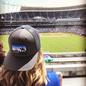 May include: A gray and black baseball cap with a blue and white patch featuring a silhouette of a person riding a horse. The cap is worn by a person sitting in a stadium.