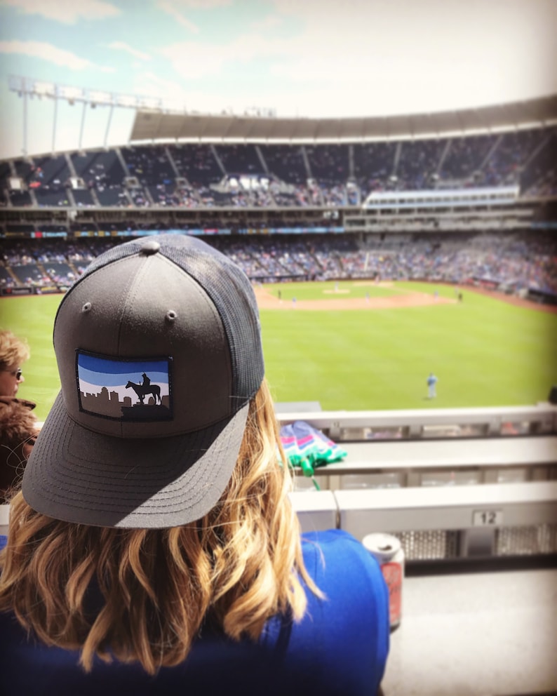 May include: A gray and black baseball cap with a blue and white patch featuring a silhouette of a person riding a horse. The cap is worn by a person sitting in a stadium.
