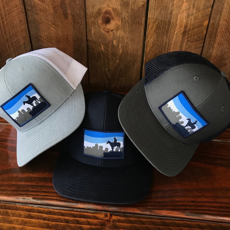 May include: Three trucker hats with a blue and white skyline patch featuring a silhouette of a person riding a horse. The hats are gray, black, and dark gray.