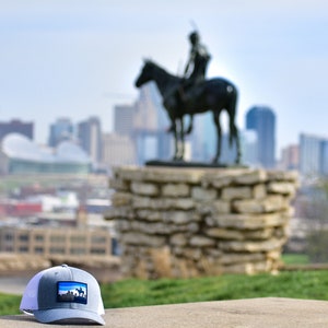 May include: A gray and white trucker hat with a blue patch featuring a cityscape. The hat is sitting on a concrete surface with a blurred background of a city skyline and a statue of a person on horseback.