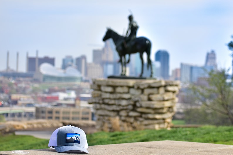 May include: A gray and white trucker hat with a blue patch featuring a cityscape. The hat is sitting on a concrete surface with a blurred background of a city skyline and a statue of a person on horseback.