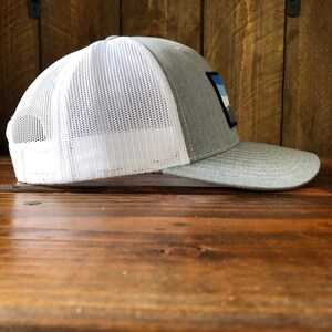 May include: A gray and white trucker hat with a blue, white, and gray patch on the front. The patch has a mountain range design.