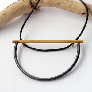 Könnte beinhalten: Eine minimalistische Halskette mit einem schwarzen Band und einem goldenen Barren-Anhänger. Der Barren hängt an einem schwarzen Metallring.