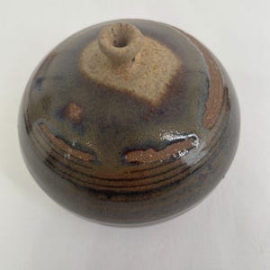 Puede incluir: Un pequeño jarrón redondo de cerámica marrón y beige con una abertura estrecha. El jarrón tiene un esmalte moteado con vetas marrones y beige.