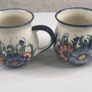 May include: Two rounded ceramic mugs with a cream-colored base, featuring a hand-painted floral pattern in shades of blue, green, and orange. Each mug has a dark blue rim and a matching dark blue handle, with a bulbous shape and a wide opening.