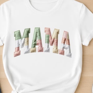 Puede incluir: Camiseta blanca con la palabra "MAMA" en letras coloridas estilo patchwork en la parte delantera. Las letras son en tonos pastel de verde, rosa, amarillo y blanco. La camiseta tiene cuello redondo.