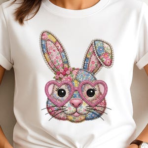 Può includere: T-shirt bianco con un disegno a forma di coniglio colorato. Il coniglio ha occhiali a forma di cuore e orecchie e viso a fantasia floreale. Il design include un bordo perlato intorno alle orecchie e un naso rosa.