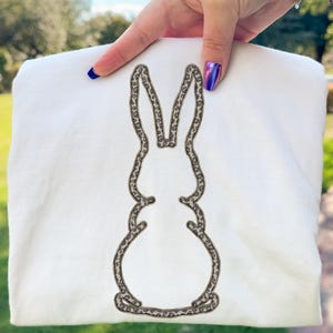 Peut inclure: Un sweat-shirt blanc plié avec un contour de lapin orné d'un motif animalier marron et beige. Le sweat-shirt est tenu par une main avec du vernis à ongles bleu et violet, sur un arrière-plan extérieur flou.