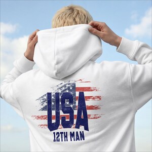 Puede incluir: Sudadera con capucha blanca con un diseño desgastado de la bandera estadounidense en la espalda. El gráfico de la bandera incluye el texto "USA" en letras azules en negrita y las palabras "12TH MAN" debajo. La sudadera tiene capucha y mangas largas.