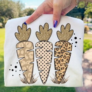 May include: A white t-shirt featuring three carrot designs with glittery gold tops and patterned bodies. The carrot bodies have a leopard print, polka dots, and a tan animal print. Black dots and grass accents complete the design.