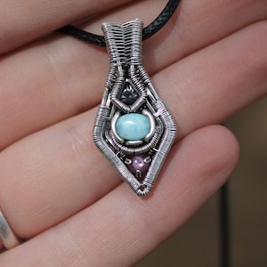 以下が含まれることがあります： 手作りのシルバーペンダントネックレス。ワイヤーラップのデザインです。ペンダントの中央には水色の楕円形の石があり、下には小さな紫色の石があります。ネックレスは黒いコードに付いています。