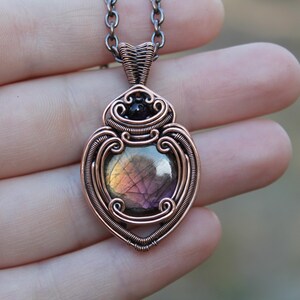 Pode incluir: Um pingente de cobre enrolado em arame com uma pedra preciosa de labradorita redonda e iridescente com reflexos roxos e dourados, acentuado por uma pequena conta preta no topo. O pingente está suspenso em uma corrente de cobre escuro.