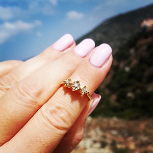 Puede incluir: Un anillo de oro con tres pequeños diamantes engastados en un diseño delicado. El anillo se lleva en un dedo con esmalte de uñas rosa claro.