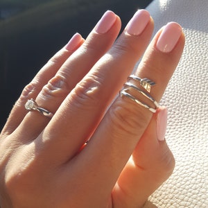 Könnte beinhalten: Eine Hand mit rosa Nagellack trägt einen silbernen Ring mit einem Pfeilmotiv am Daumen. Der Ring sitzt auf dem oberen Knöchel des Daumens. Die Hand trägt auch einen silbernen Ring mit einem Diamanten am Ringfinger.