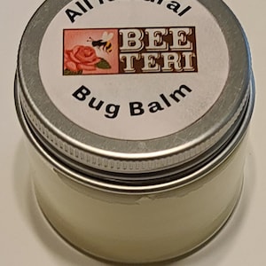 May include: A small, round metal tin of "All Natural Bee Teri Bug Balm." The label features a pink floral design with a bee. The balm is a light yellow color, and the lid is silver.