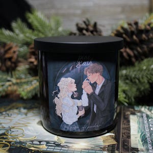 Puede incluir: Vela negra con tapa, con una ilustración de una pareja. La obra de arte representa a un hombre y una mujer en un ambiente romántico. La vela está etiquetada como "Pine & Ash" con texto adicional.